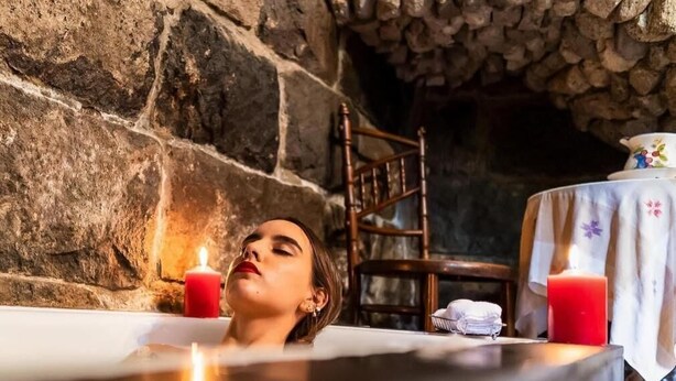 A person relaxing in a bathtub with candles, a wooden chair, and a table with a cloth and a teapot in a stone-walled room. - Hacienda Hato Verde (Latacunga)