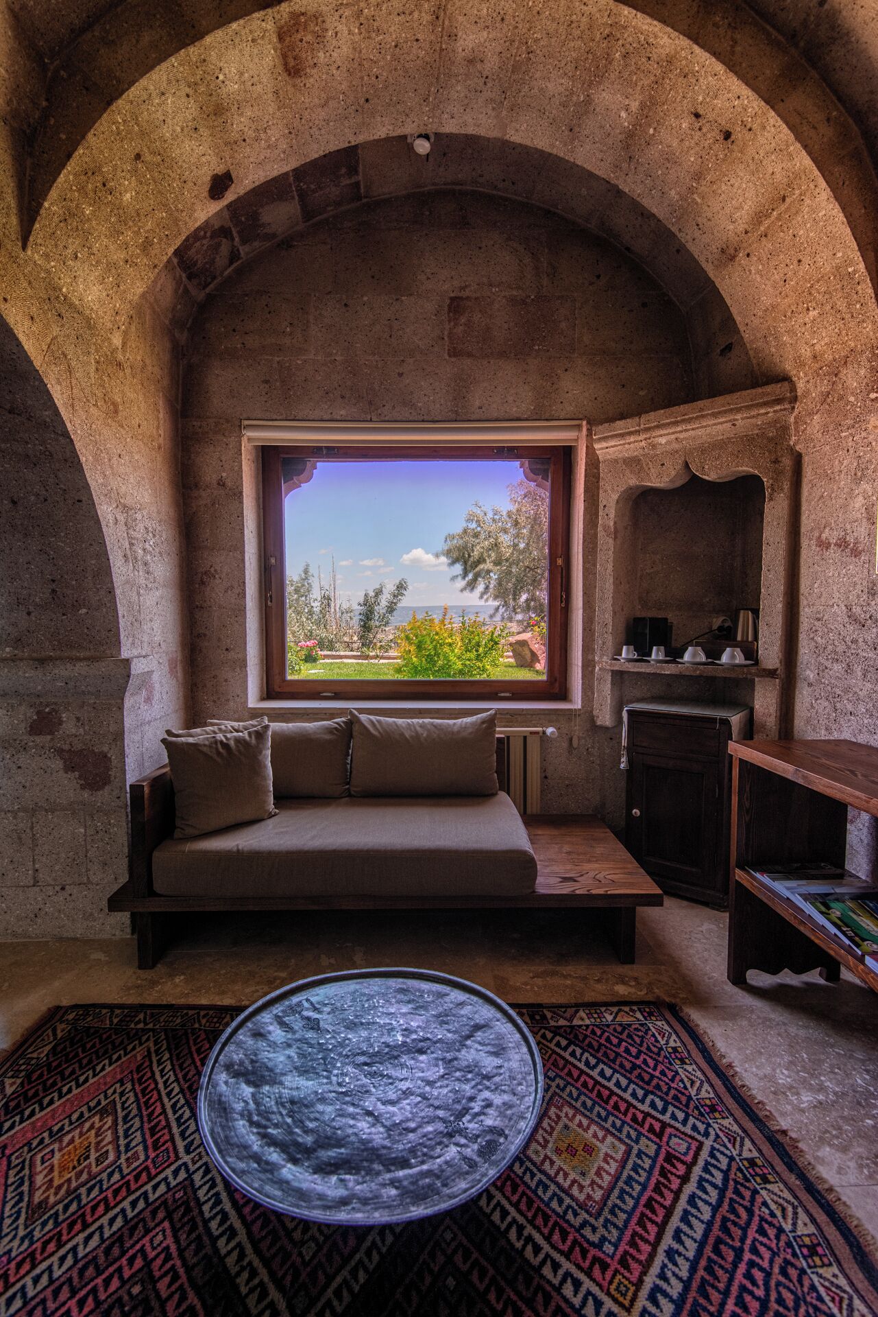 Une pièce accueillante avec une arche en pierre, un canapé agrémenté de coussins, une petite table et, par la fenêtre, une vue sur la verdure et une étendue d’eau.