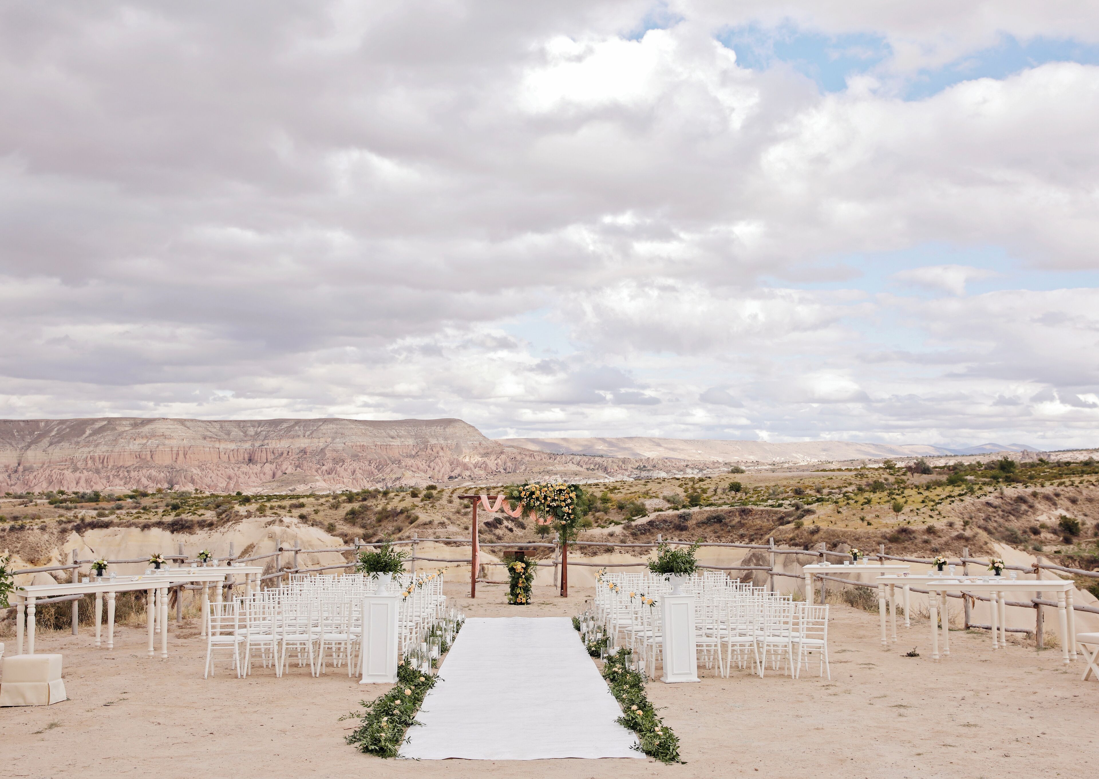 Un lieu de cérémonie de mariage en plein air, avec une allée et des chaises blanches, situé dans un paysage désertique et dominé par des montagnes en arrière-plan.