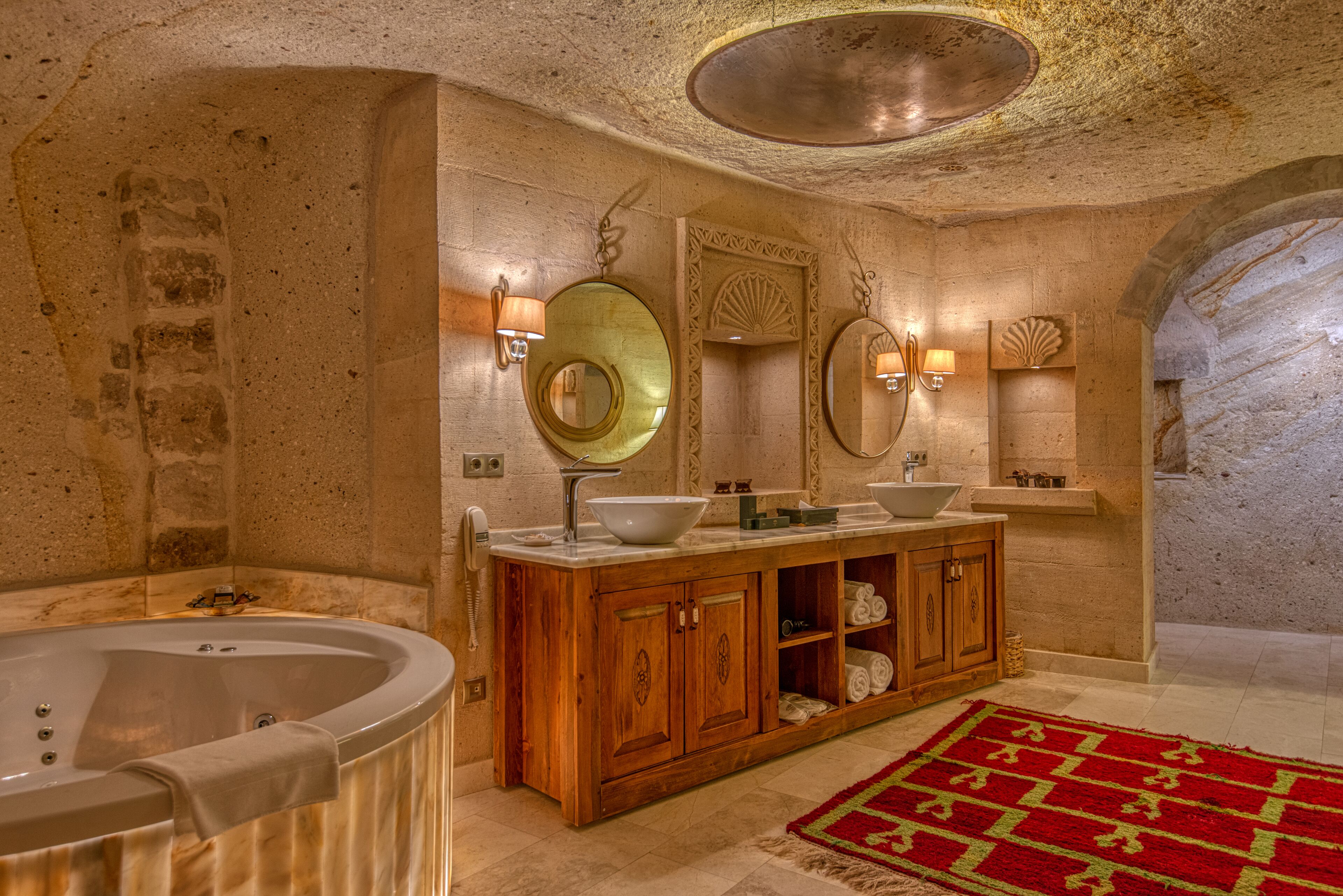 Une salle de bain luxueuse avec une baignoire îlot, un meuble-lavabo en bois doté d’un grand miroir et un tapis rouge à motifs.
