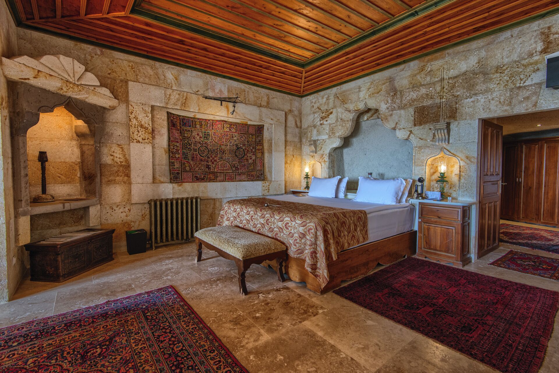 Un dormitorio con pared de piedra, techo de madera, cama con colcha estampada, un banco, mesita de noche con lámpara y un tapiz grande.
