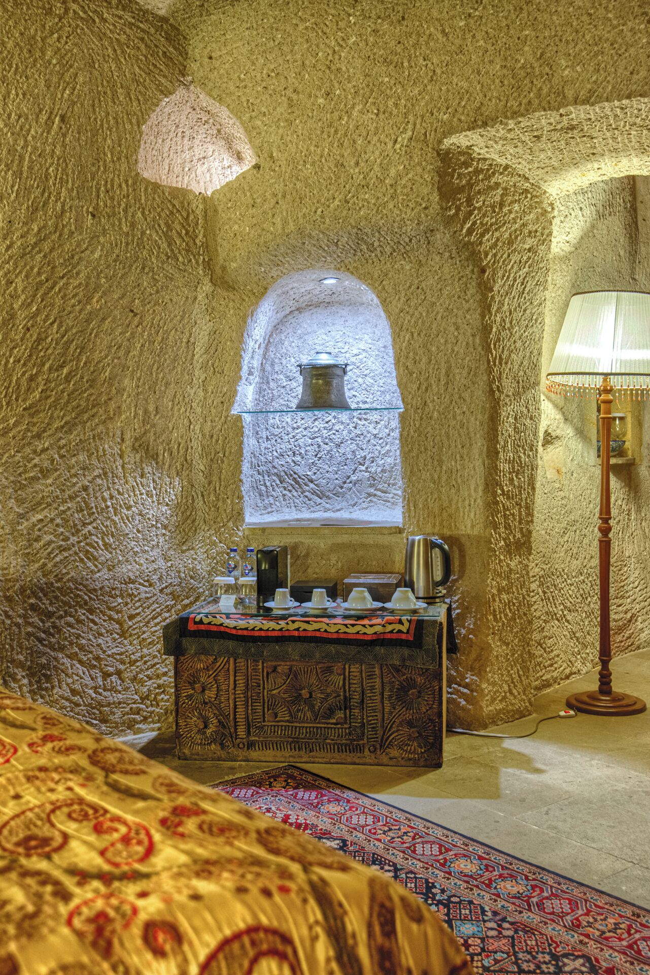 A room with a stone wall, a small window with a decorative glass, a bed with a patterned bedspread, a wooden nightstand with a lamp, and a coffee table with a tray of cups and a kettle.