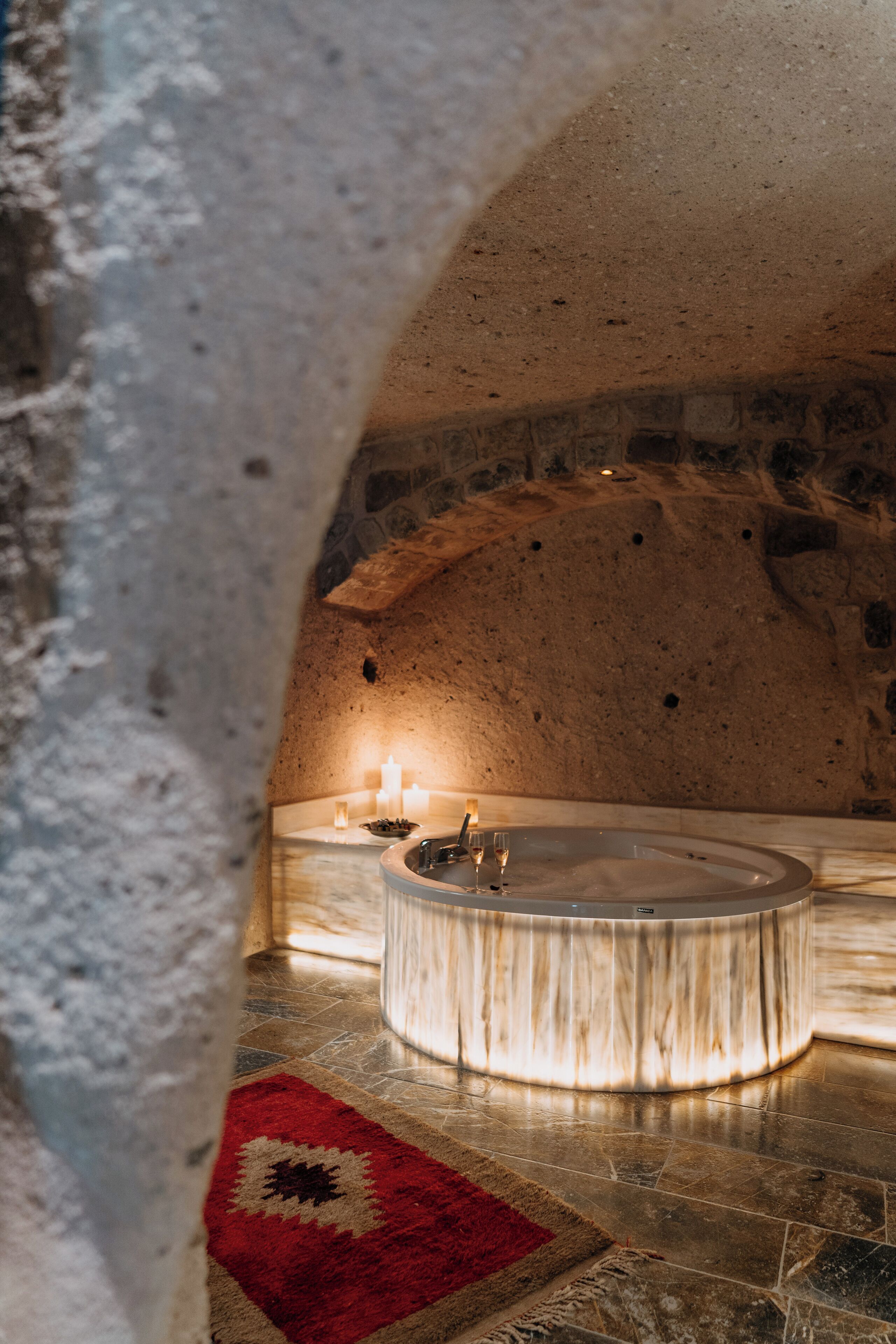 Un baño de paredes de piedra con una bañera de mármol, velas y una alfombra estampada.