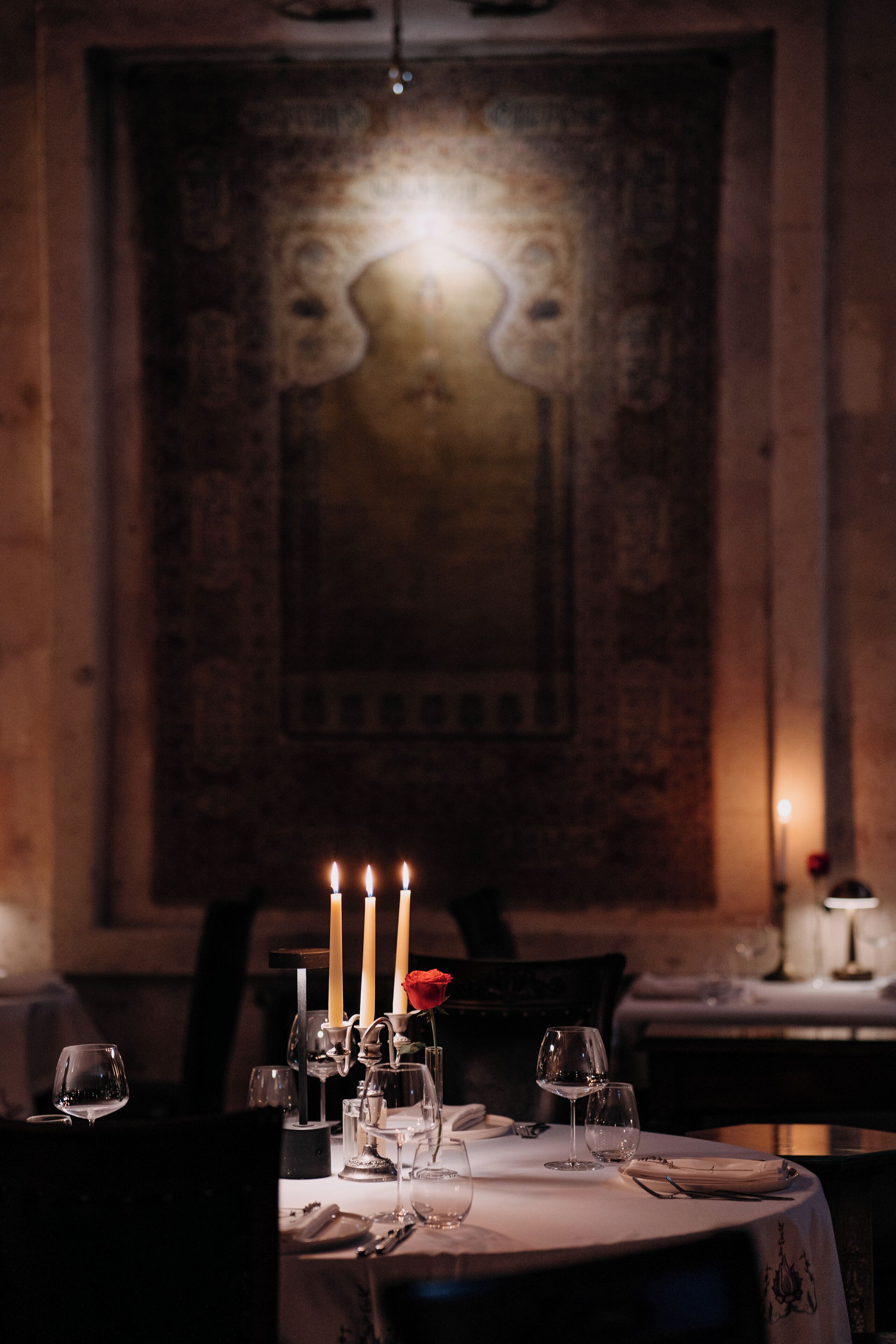 Un comedor con poca luz, una mesa preparada para dos personas, velas y una rosa.