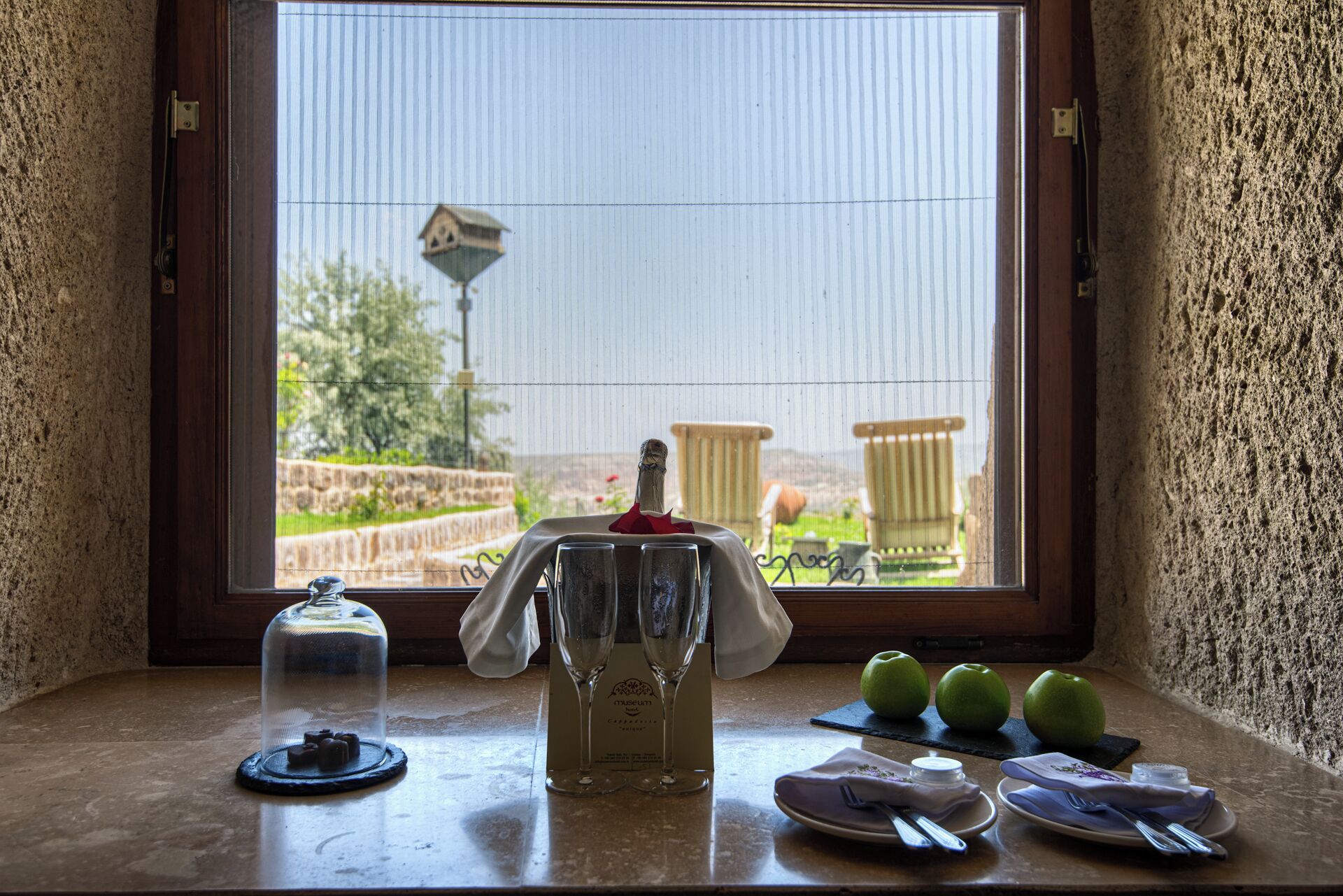 Una mesa con una botella de champán, dos copas, un vaso de agua y tres manzanas verdes sobre una encimera de piedra.