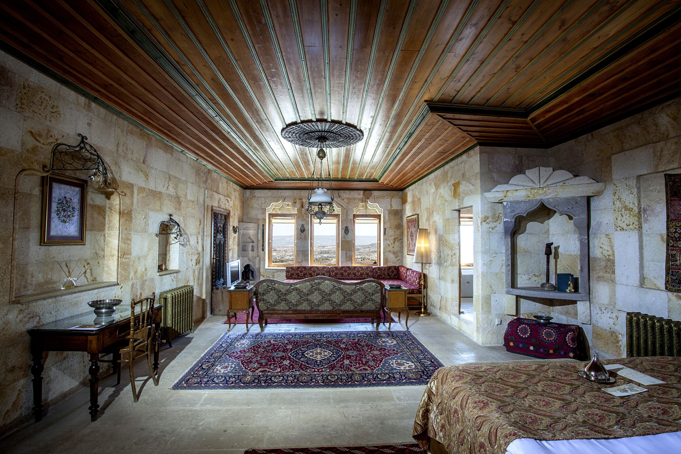 A room with a wooden ceiling, a large bed with a patterned bedspread, a sofa, a dining table with chairs, and a large rug.