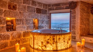 Habitación de paredes de piedra con una bañera grande iluminada, velas y una ventana que ofrece vistas de la ciudad.