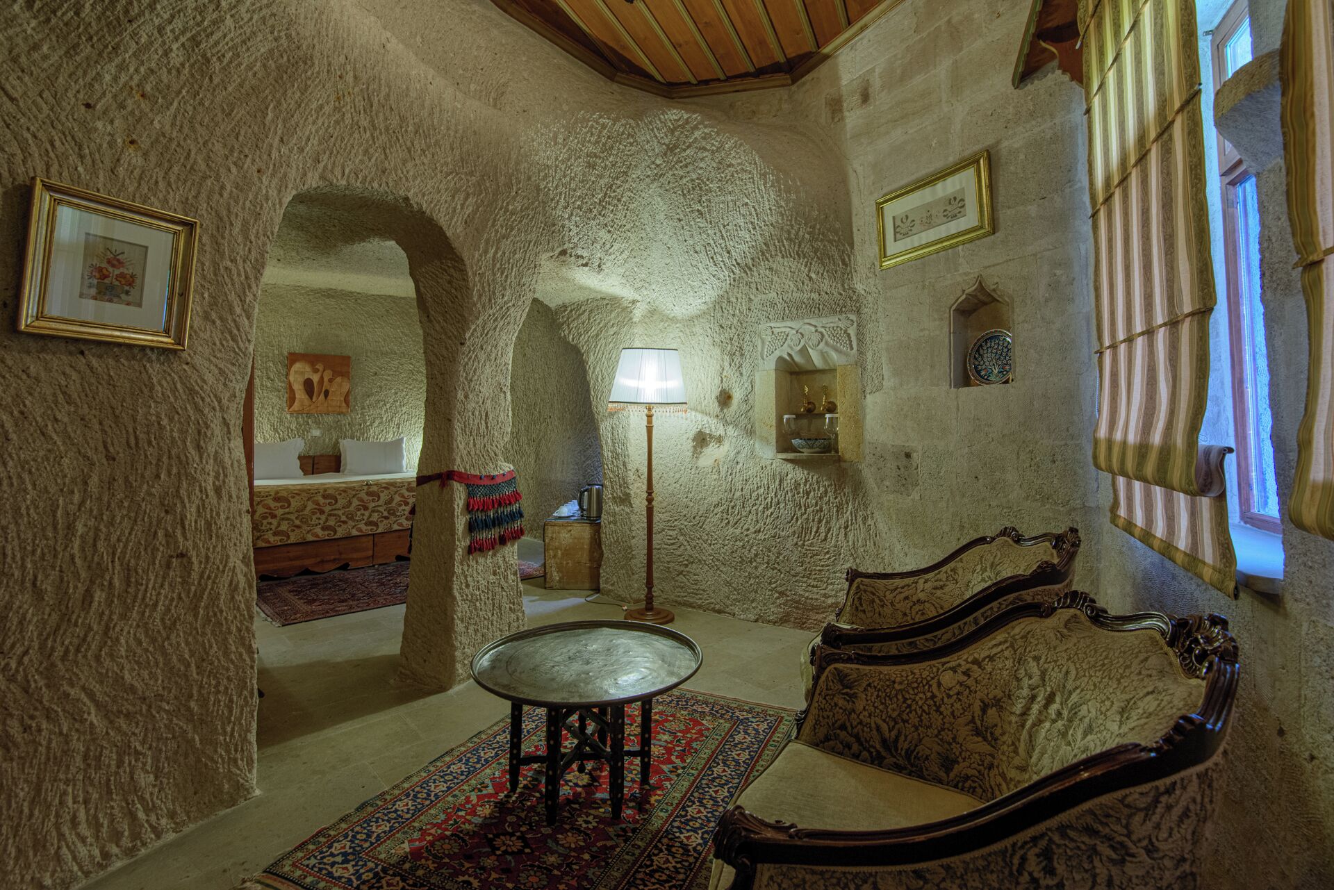 Une pièce avec un mur en pierre, un lit, une table et un canapé. Des tableaux encadrés ornent le mur et une lampe se trouve sur la table.