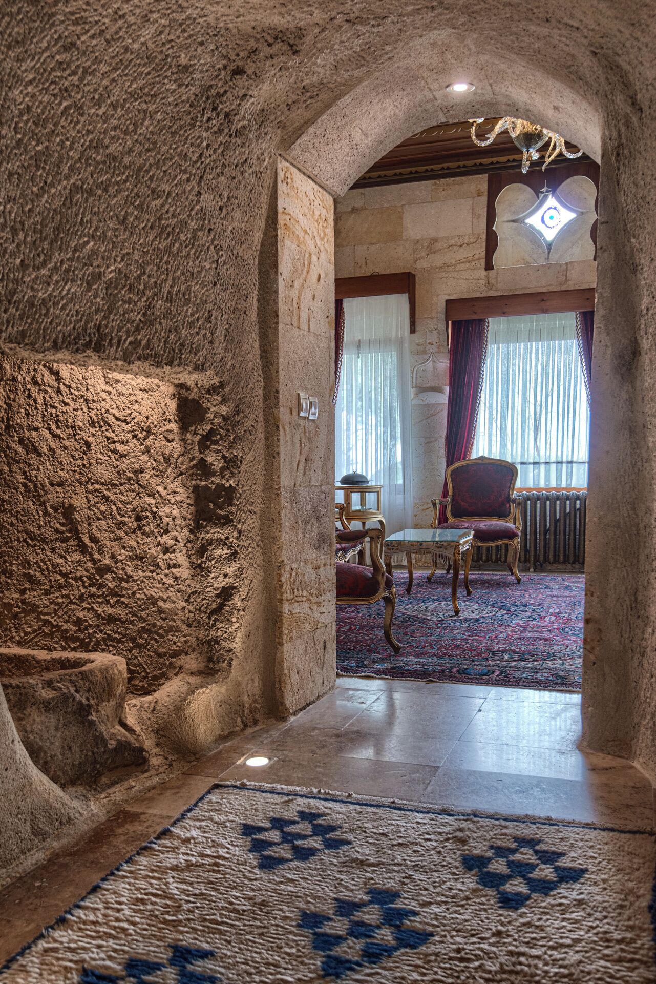 Un pasillo con paredes de piedra que conduce a una habitación con alfombra estampada, un candelabro y una ventana con cortinas.
