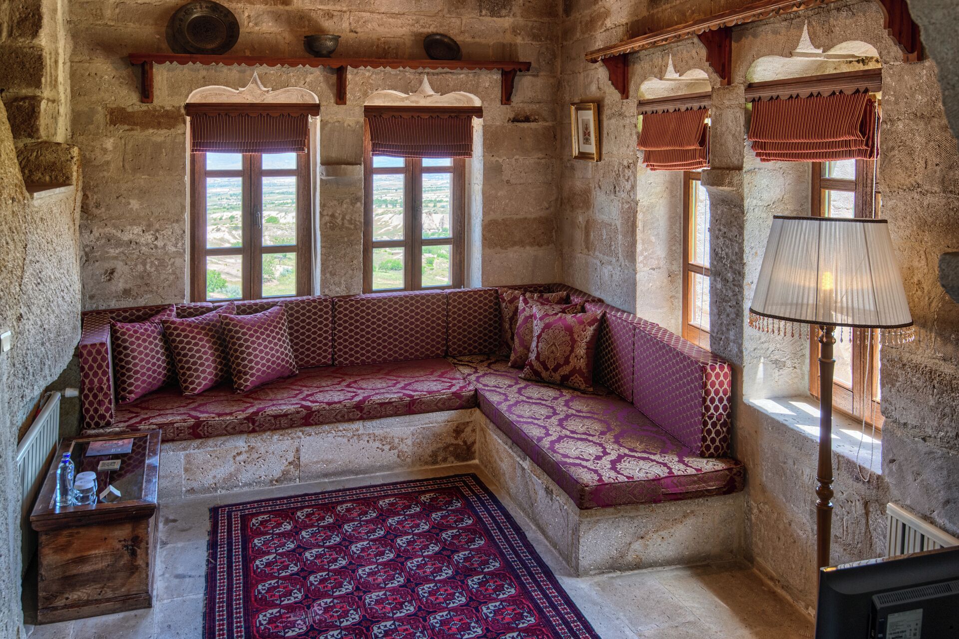 A traditional room with a stone wall, a wooden bench with patterned cushions, a red rug, and a lamp.