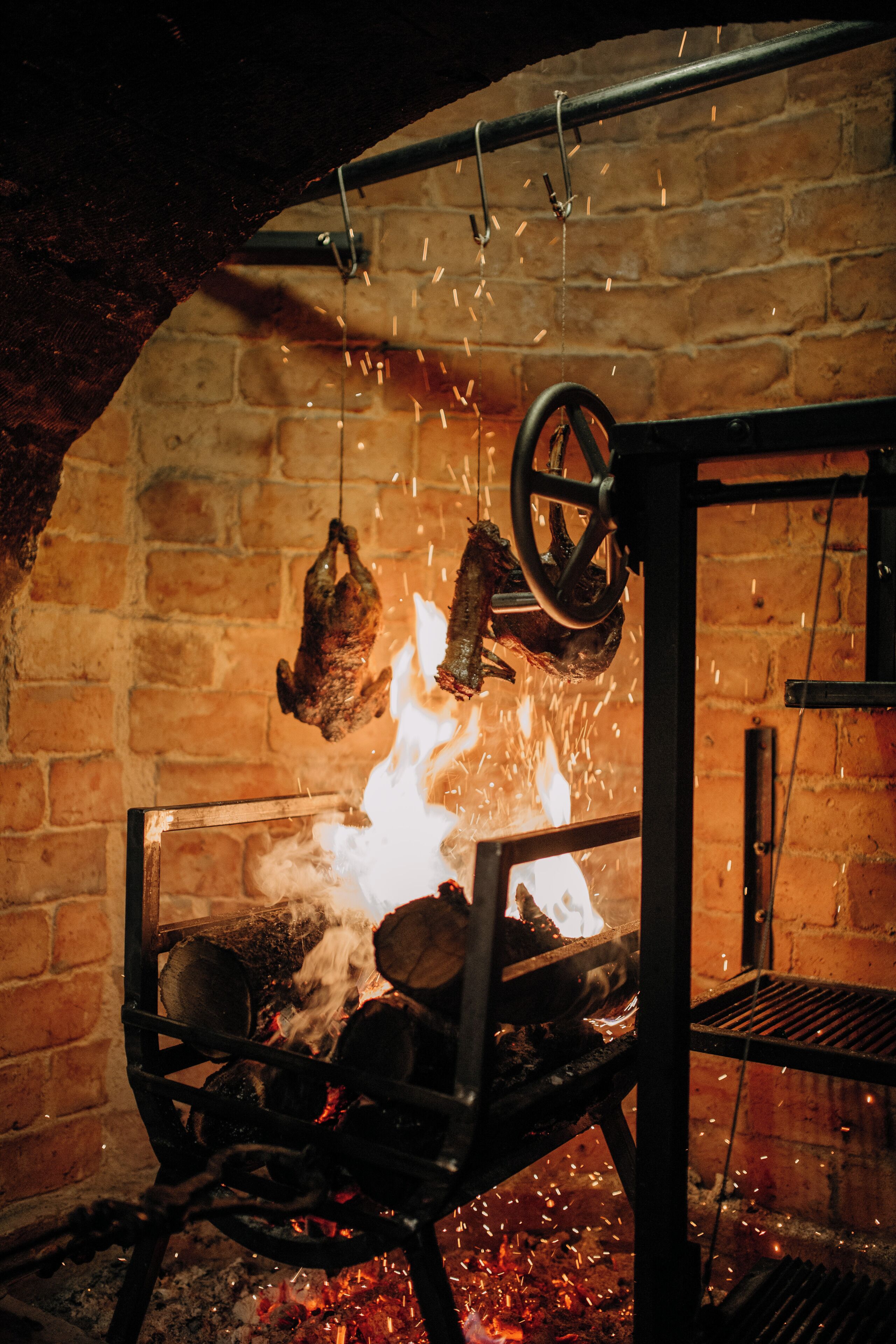 Een traditionele bakoven met een hangende vleesrek, hout brandend van binnen en zichtbare vonken.