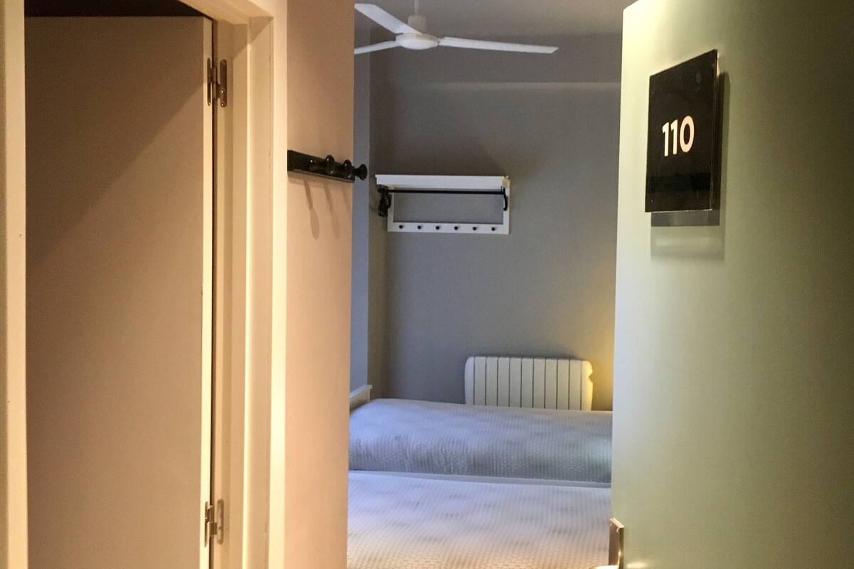 Habitación de hotel con una cama, ventilador de techo, radiador y un cartel que indica el número de habitación 110.