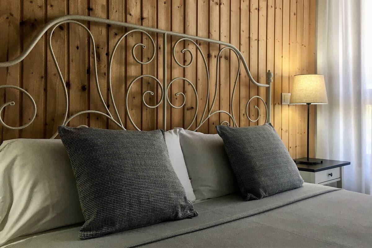 Una cama con cabecera de metal, dos almohadas, una mesita de noche con lámpara y una ventana con cortinas translúcidas.