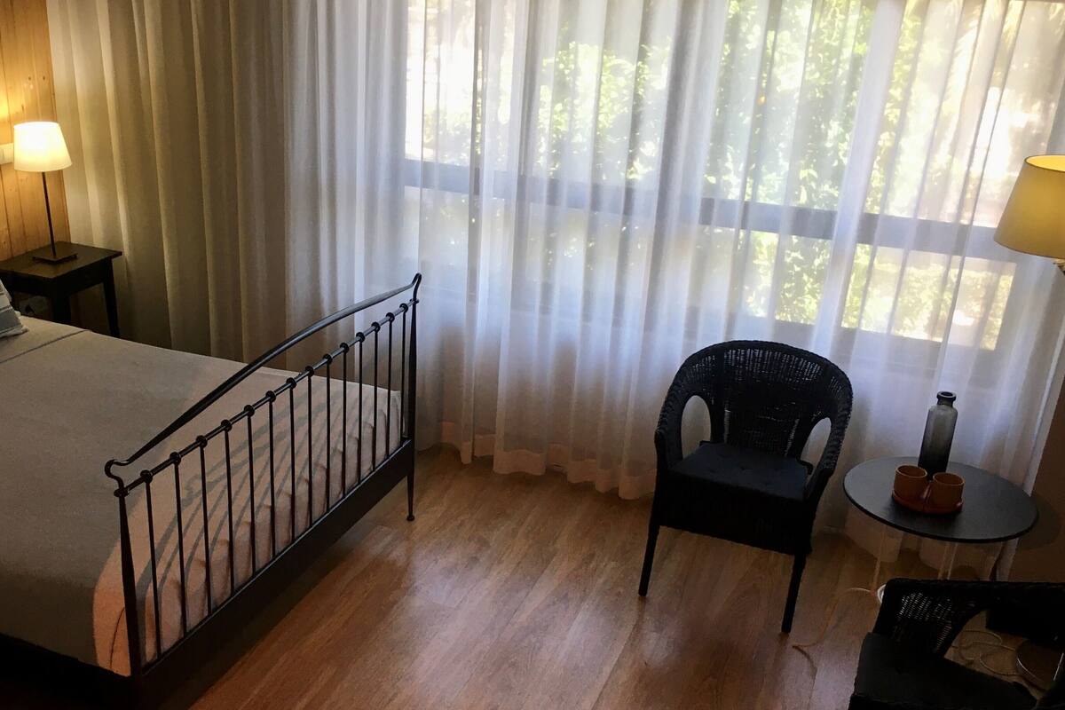 Un dormitorio con cama, una silla, una mesita de noche con lámpara y una ventana con cortinas de gasa.