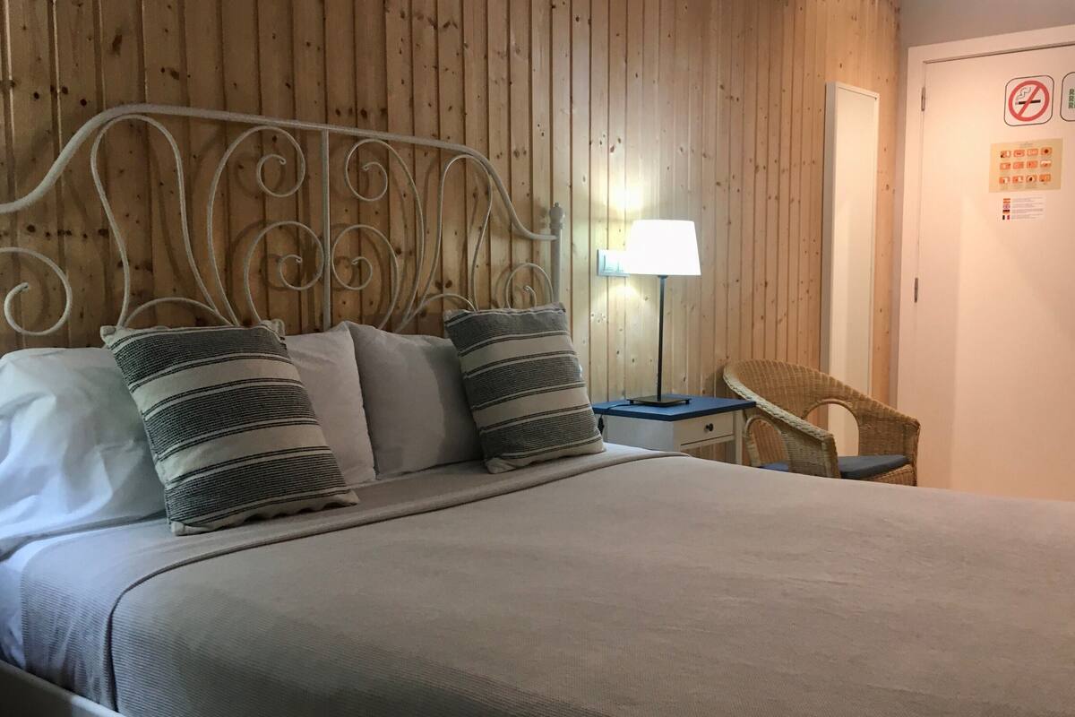 Una cama bien hecha con cabecera de metal, una mesita de noche con lámpara y una silla de mimbre.