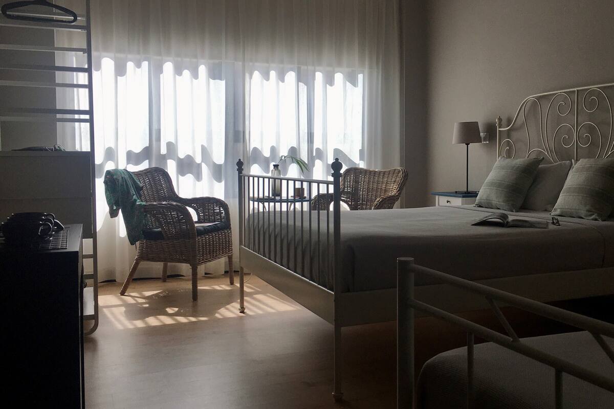 Un dormitorio con cama, una silla, una mesita de noche y una ventana con cortinas.