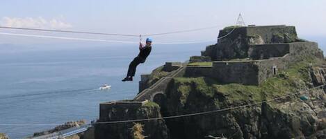 En person suser ned på en kystklip på en zip-line, med udsigt til havet og en historisk bygning i baggrunden.