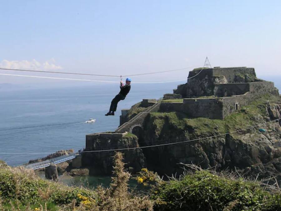 A person zip-lining over a coastal cliff with a view of the ocean and a historical building in the background.