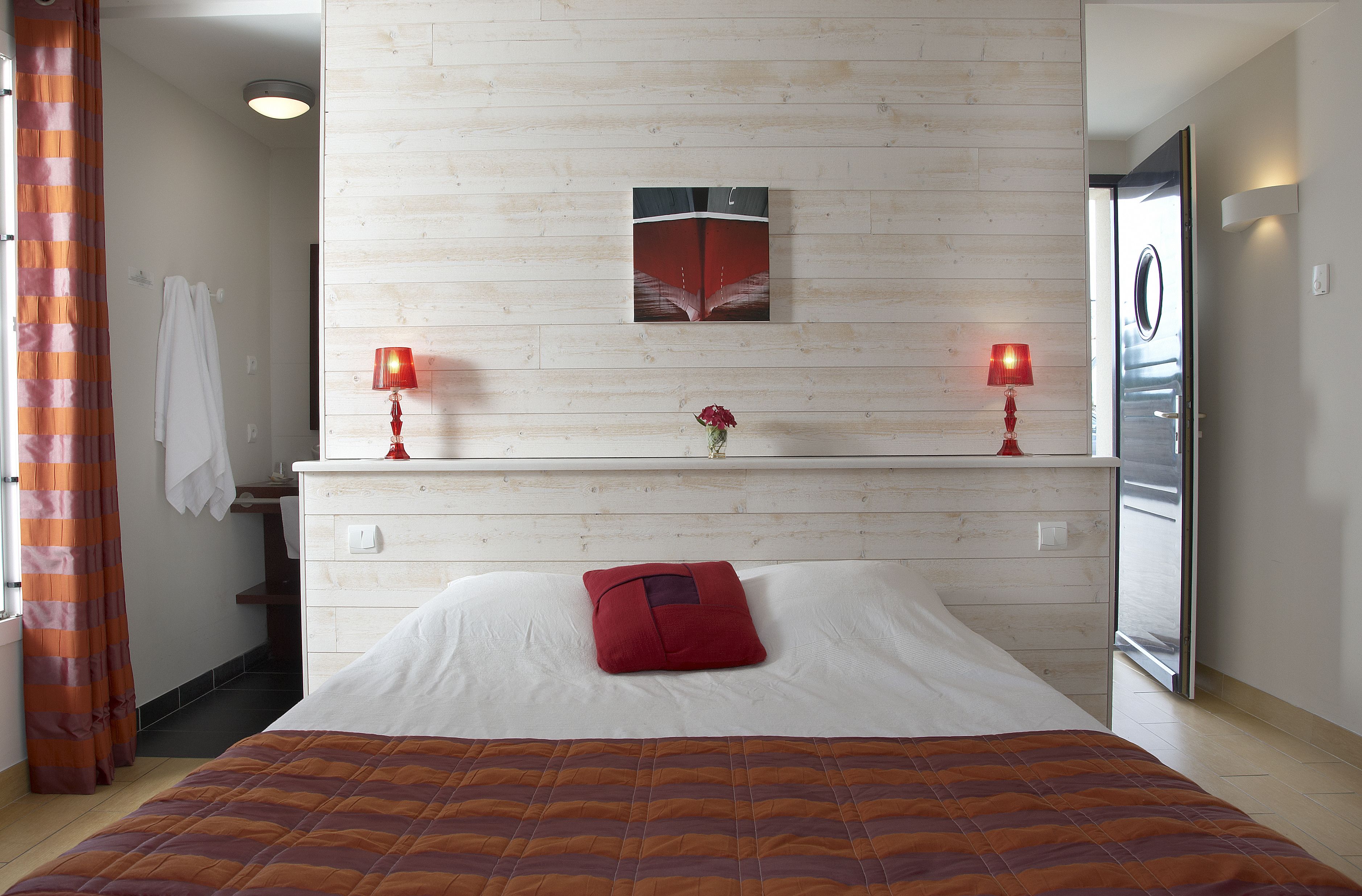 Habitación de hotel con cama, mesita de noche, espejo, lámpara roja y un cuadro en la pared.