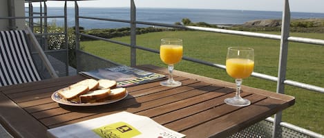 Une table en bois, avec un magazine, deux verres de jus d’orange et une assiette de sandwichs, sur un balcon donnant sur la mer.