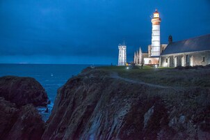 Un edificio costero con un faro, una torre y un área verde con un camino que lleva al mar.