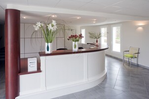 A reception area with a curved reception desk, a sign reading "bonjour facteur", and three vases with flowers.