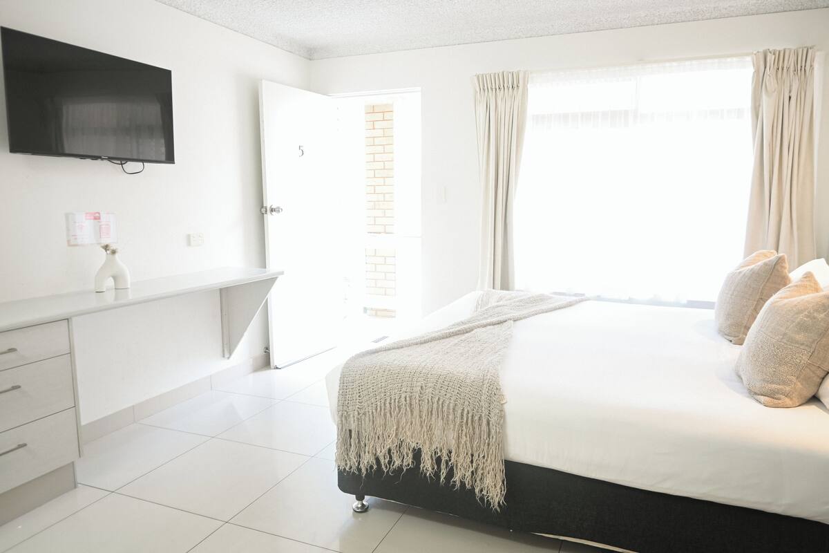 Un dormitorio con cama, televisor montado en la pared, una puerta y una ventana con cortinas.