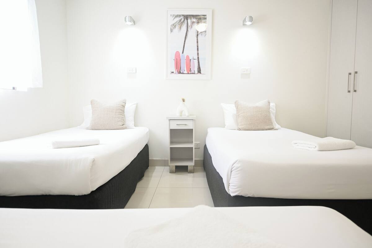 Habitación de hotel con dos camas, una mesita de noche, una pequeña mesa y un cuadro colgado en la pared.