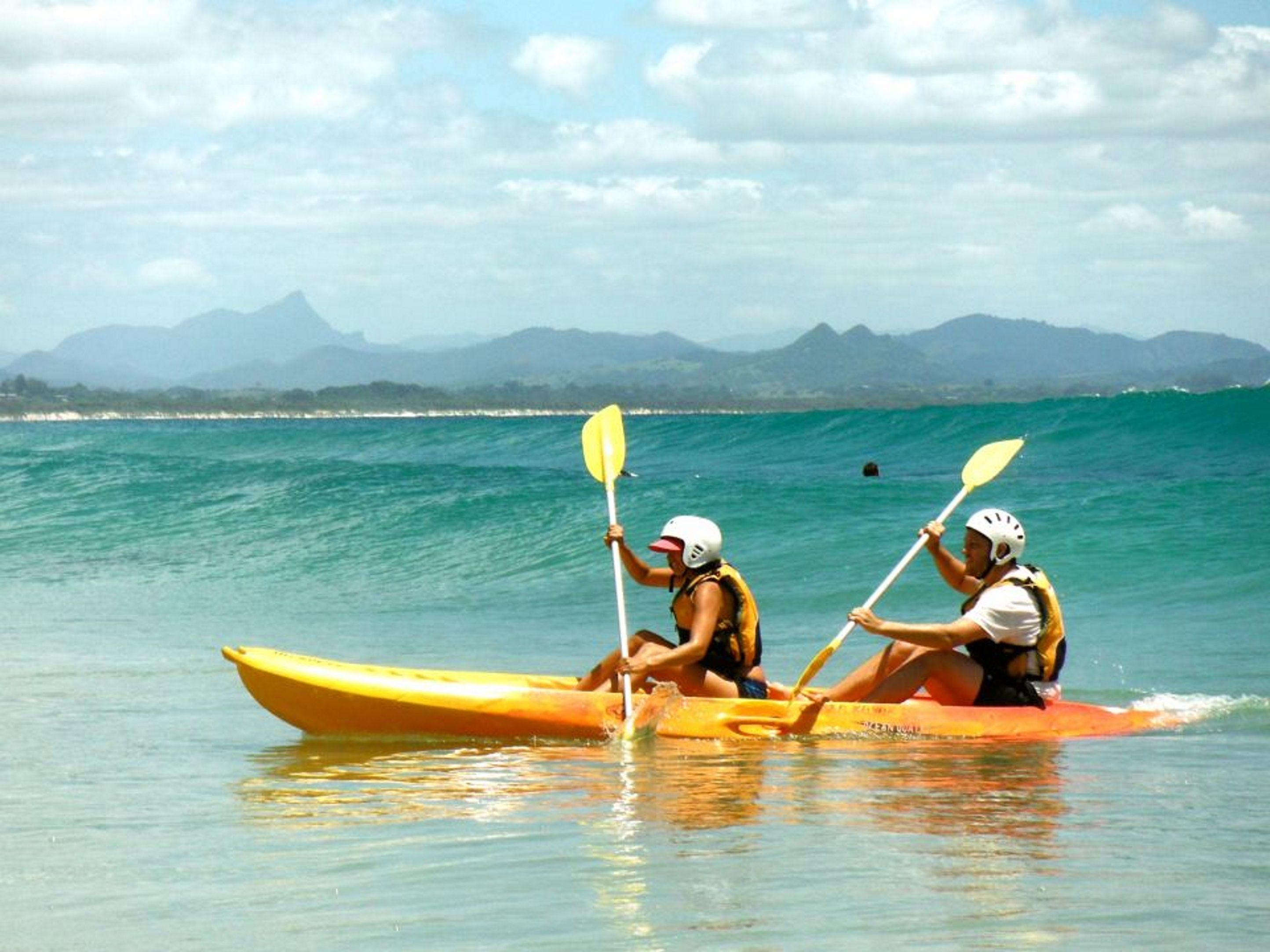 Two people kayaking on a calm sea with mountains in the background.