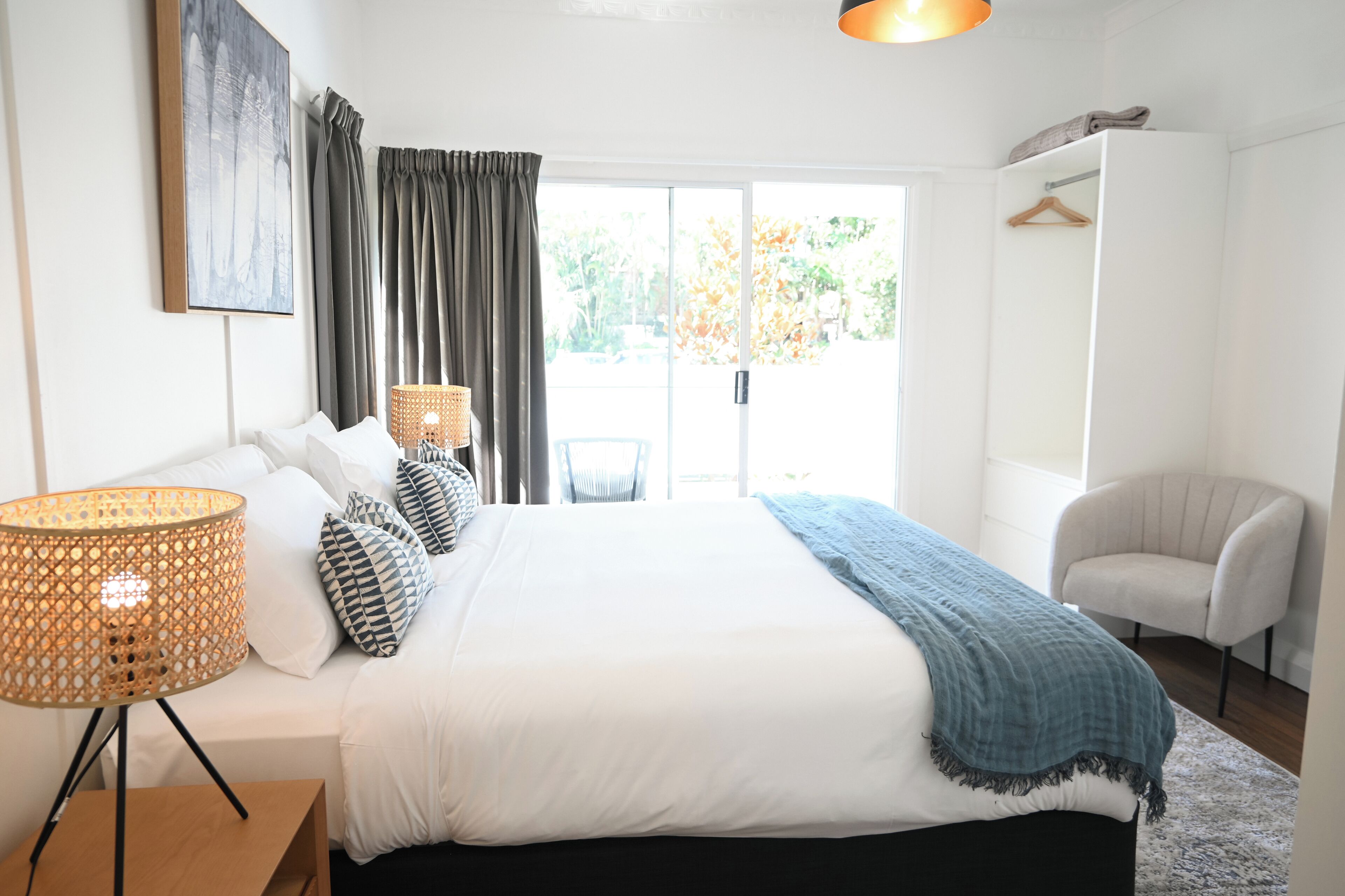 A bedroom with a large bed, a bedside table with a lamp, a chair, a wardrobe, and a view of the outdoors.