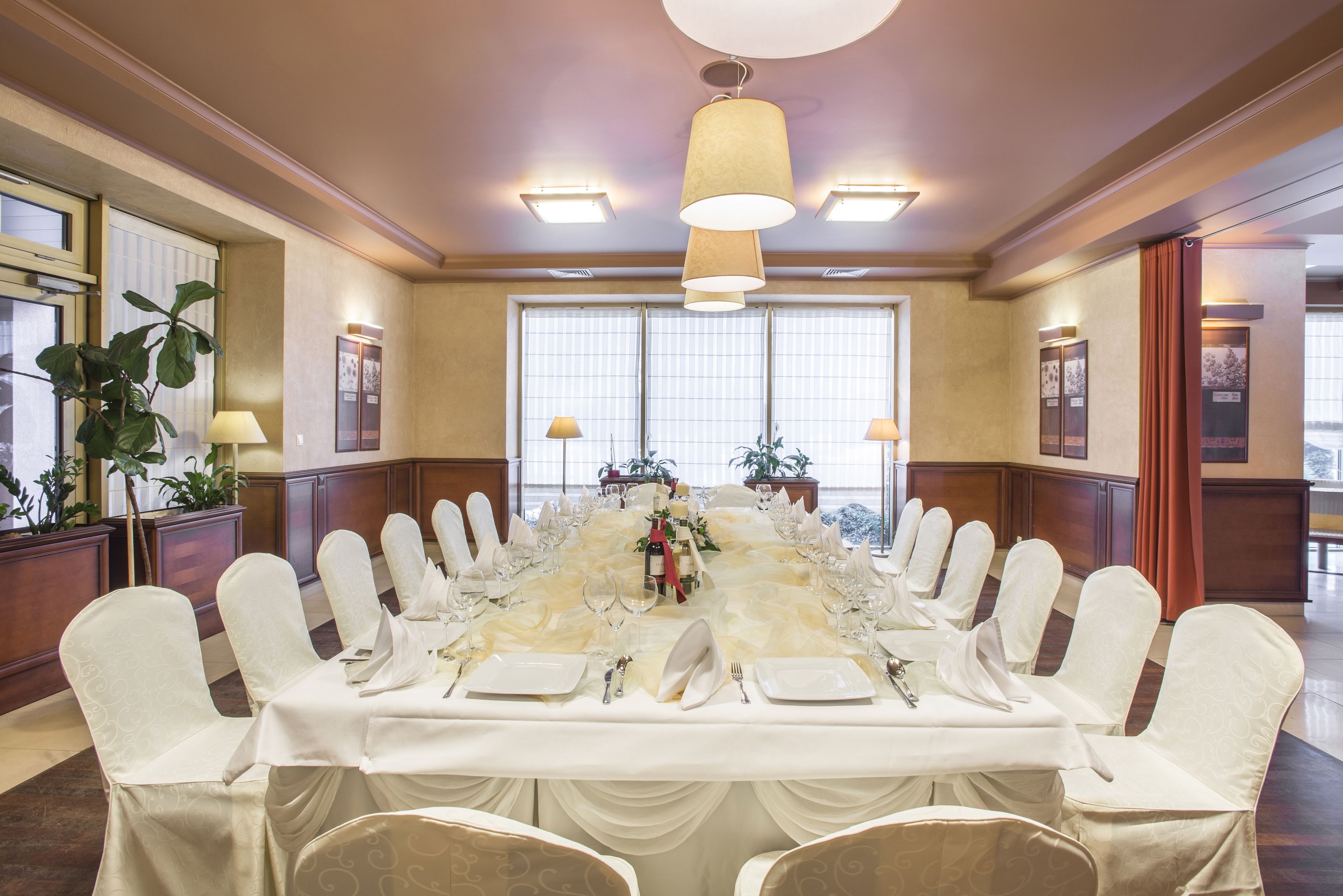 A conference room with a long table set for a formal event, surrounded by chairs with white covers, and large windows with blinds.