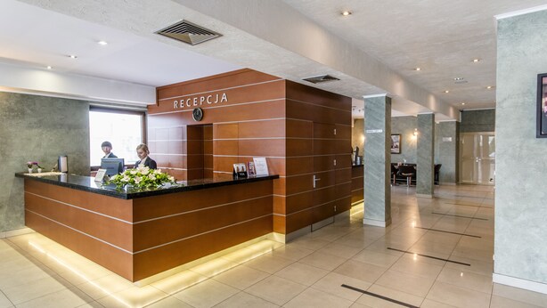 A reception area with a wooden reception desk, a receptionist, and a sign that reads "RECEPCJA". - Business Hotel Vega (Wroclaw)
