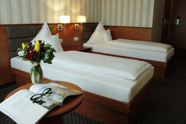 Habitación de hotel con dos camas individuales, una mesita de noche con un jarrón de flores, una mesa con una revista y vasos, y dos lámparas de pared.