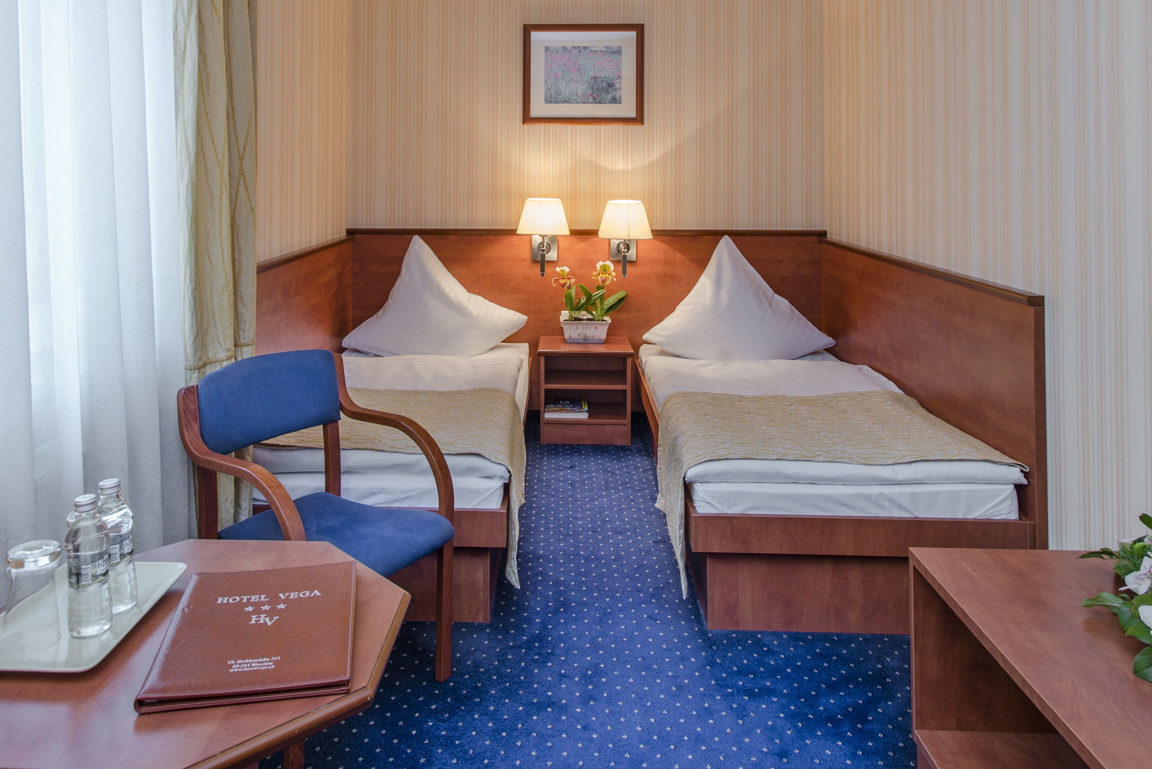 Hotelkamer met twee enkele bedden, een houten nachtkastje met lampen, een stoel, een klein tafeltje met een dienblad vol waterflessen en een blauw tapijt.