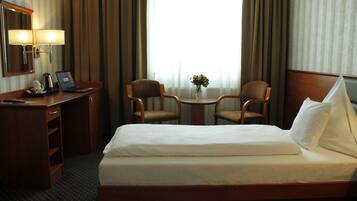 A hotel room with a bed, a desk with a laptop, two chairs, a small table with a vase of flowers, and a window with curtains.