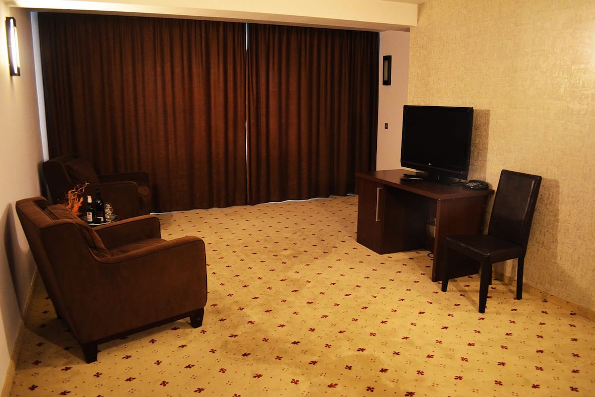 A hotel room with a large window, a flat-screen TV, a sofa, a chair, and a small table with a bottle and glasses.