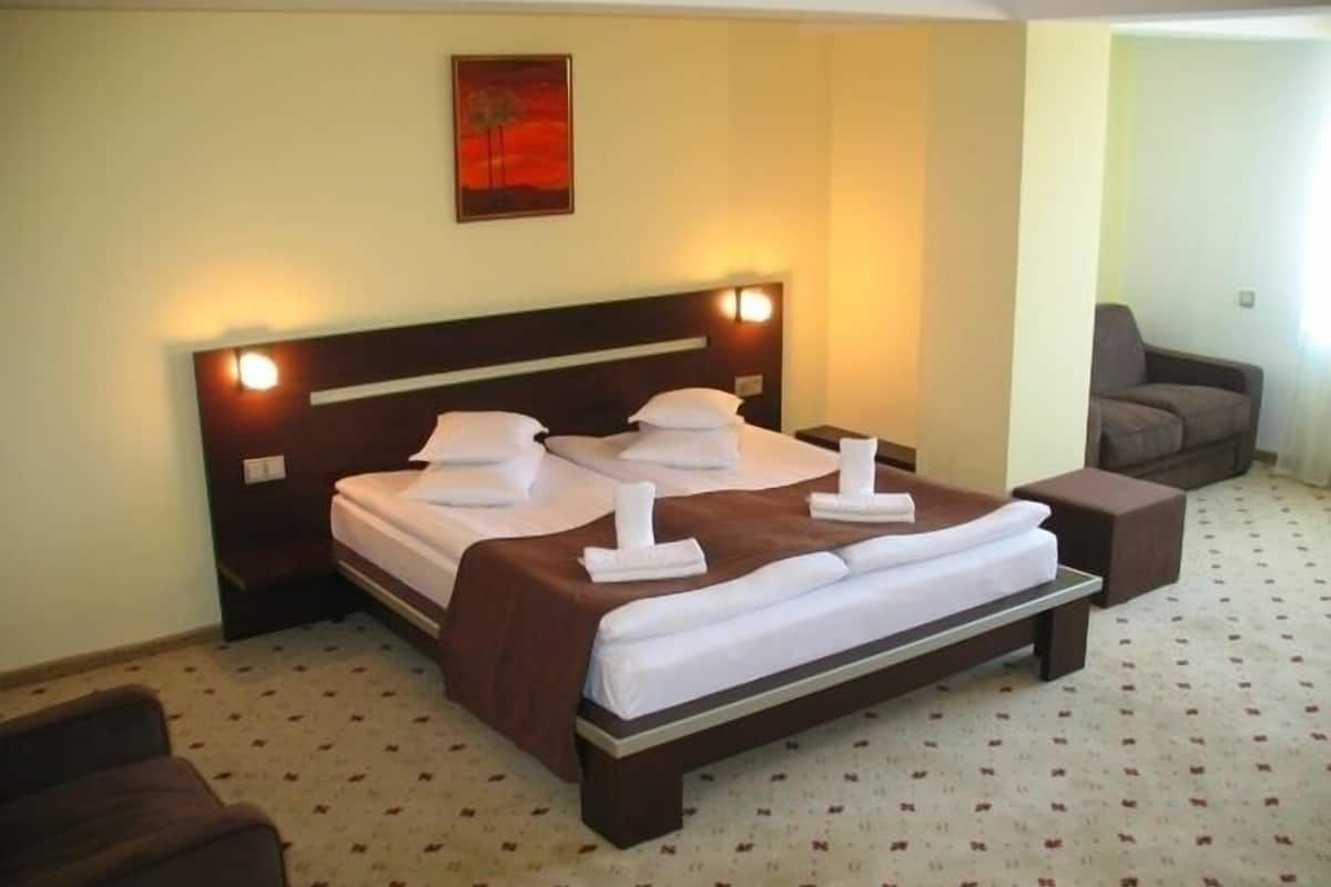 A neatly made double bed with a brown headboard and white linens, a small bedside table, and a brown armchair.
