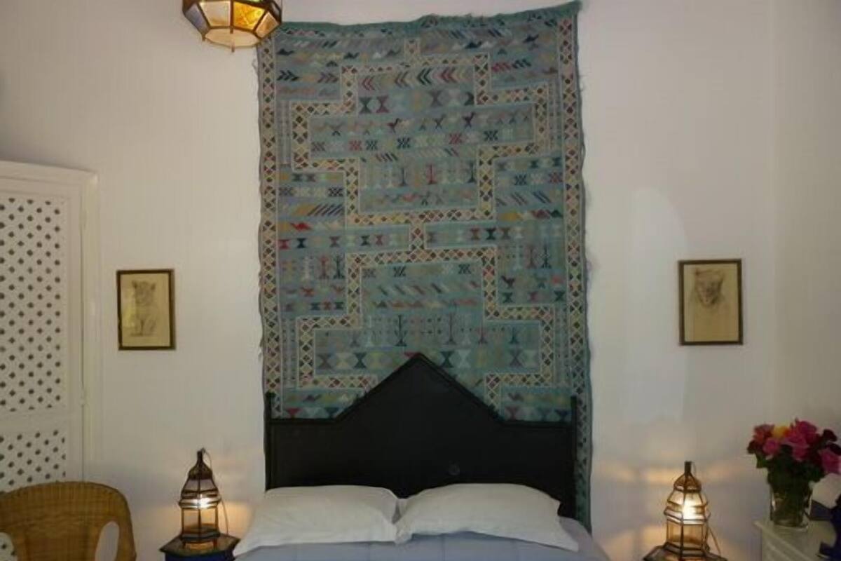 A bedroom with a patterned tapestry, a bed with white linens, two wall-mounted lanterns, and a wicker chair.
