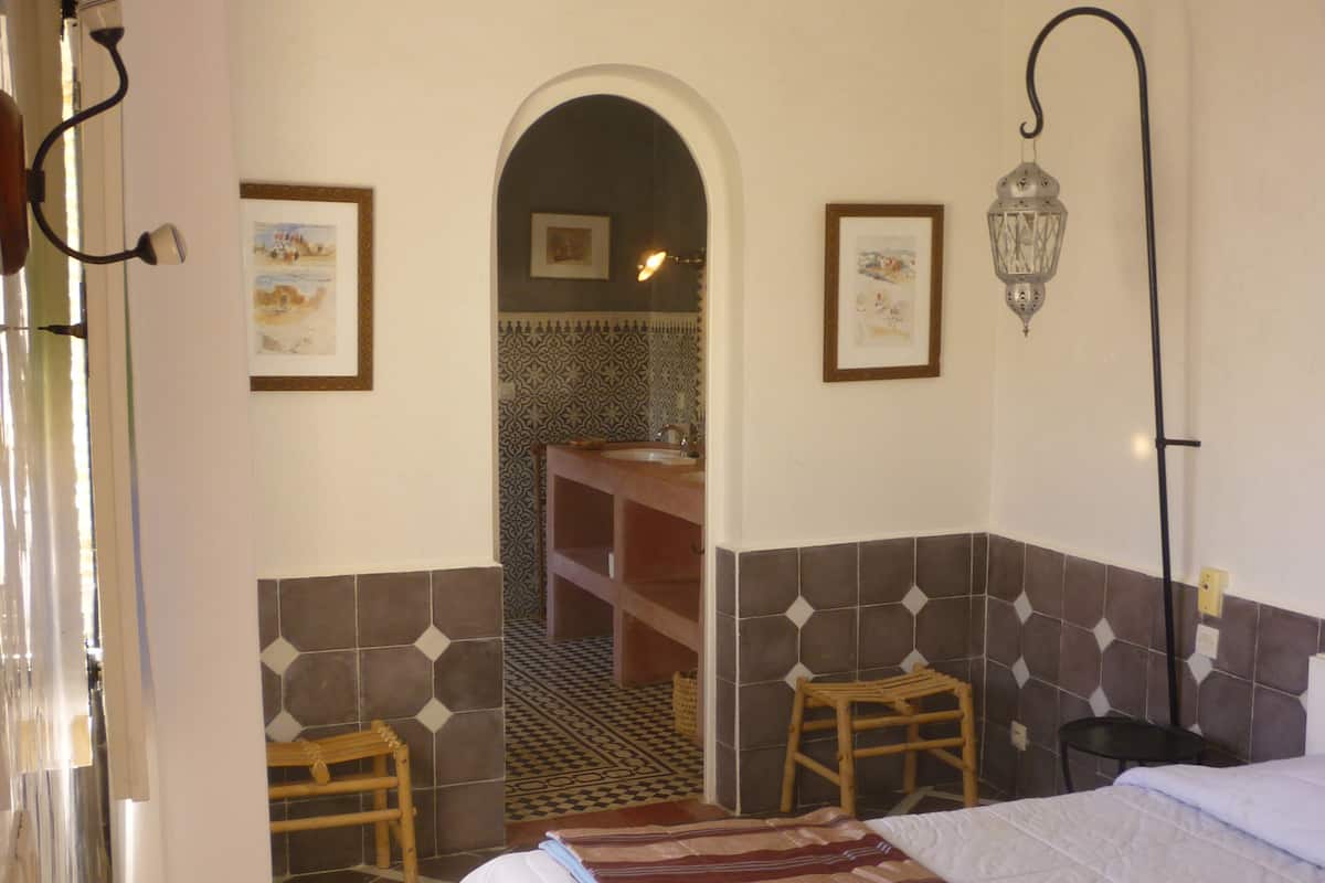 A hotel room with a bed, a wooden stool, a small table, a wall-mounted lamp, and a picture frame.