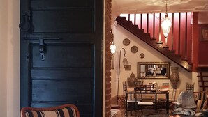 Una habitación con una puerta negra grande, un sillón estampado, un comedor con una araña, y una escalera que sube a otro nivel.