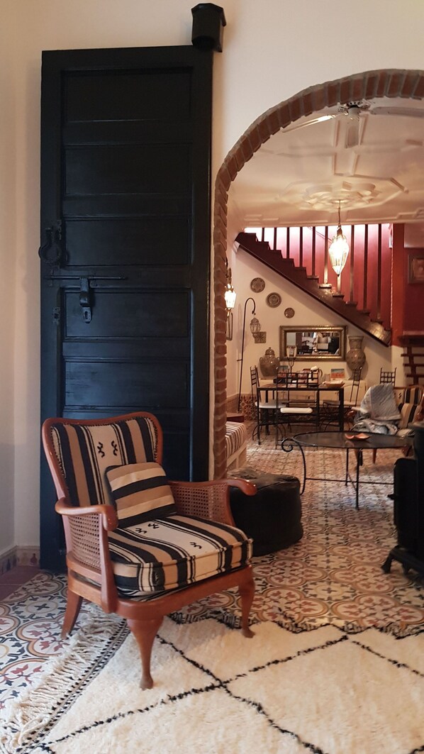 Una habitación con una puerta negra grande, un sillón estampado, un comedor con una araña, y una escalera que sube a otro nivel.