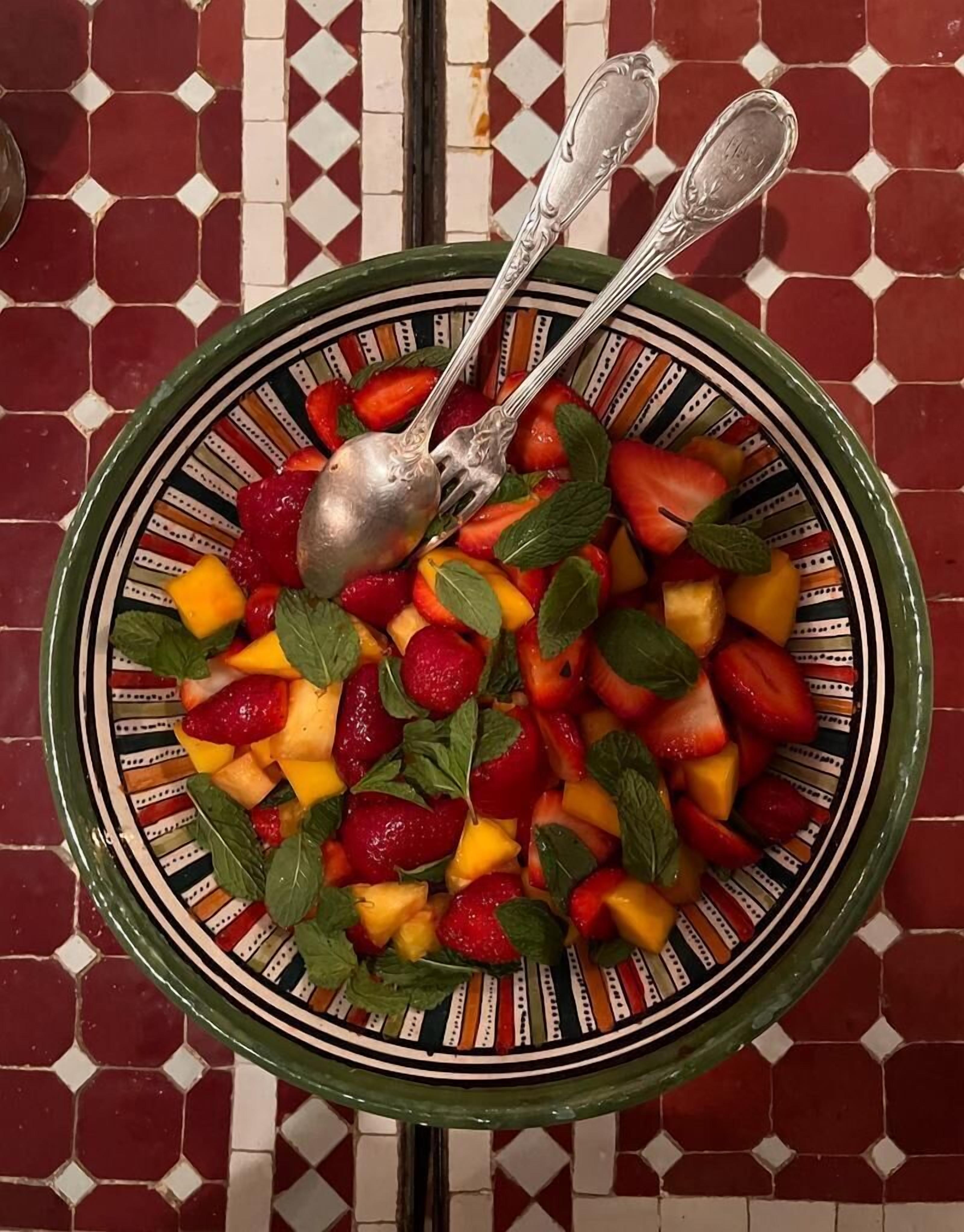 En skål med fruktsallad, garnerad med myntablad, och två skedar på en tallrik med mönster.