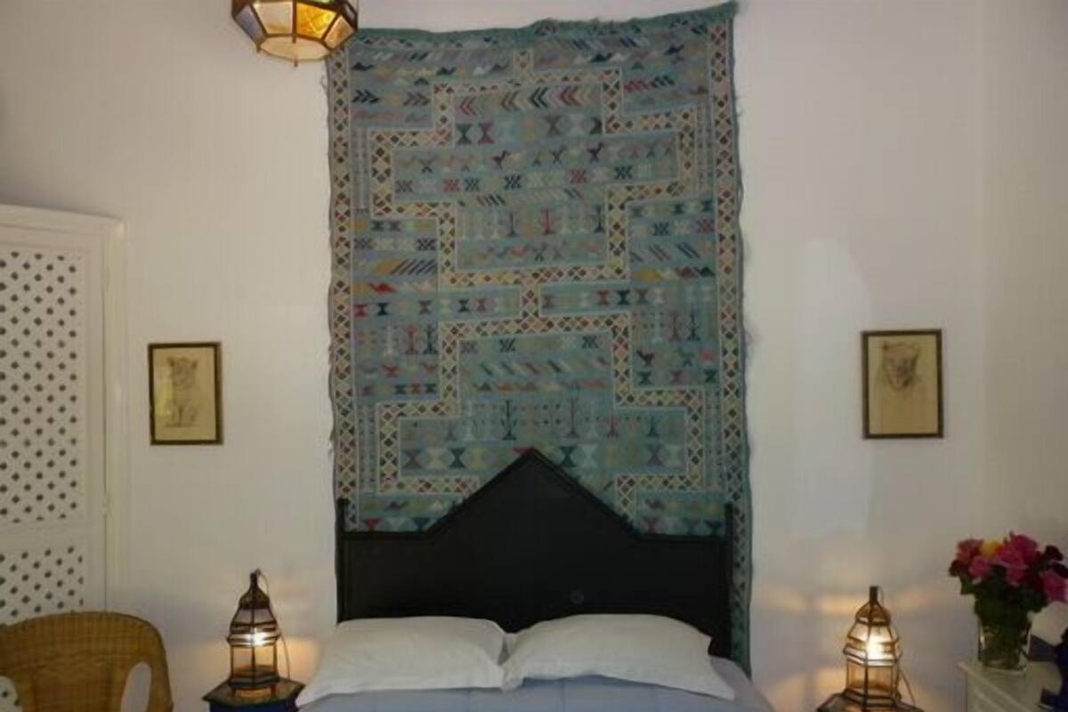 A bedroom with a patterned tapestry, a bed with white linens, two wall-mounted lanterns, and a chair.