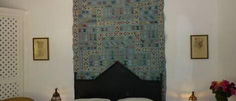 Un dormitorio con un tapiz estampado, una cama con ropa blanca, dos faroles fijados a la pared y una silla.