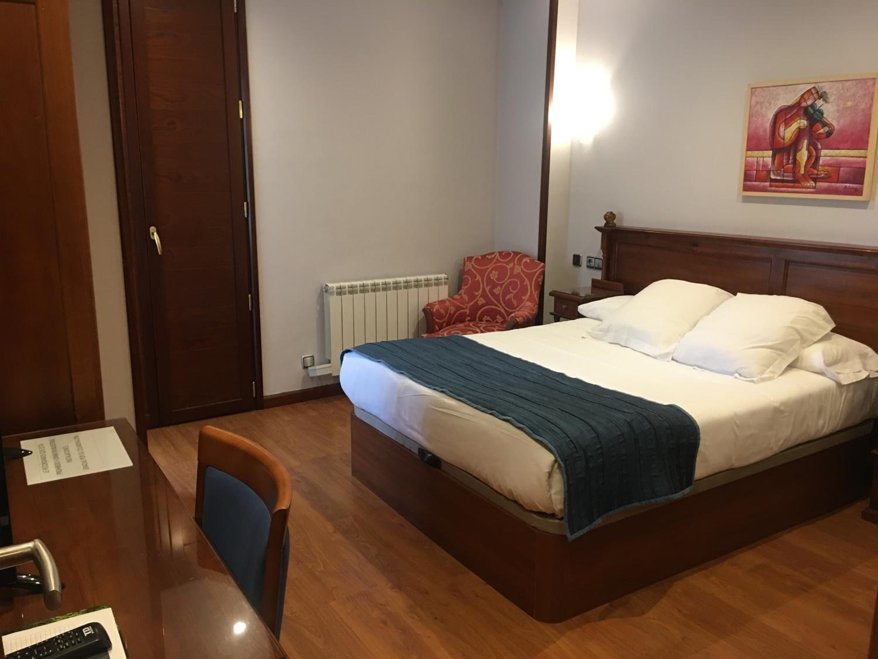 A hotel room with a wooden bed, a desk with a chair, a red patterned armchair, and a painting on the wall.