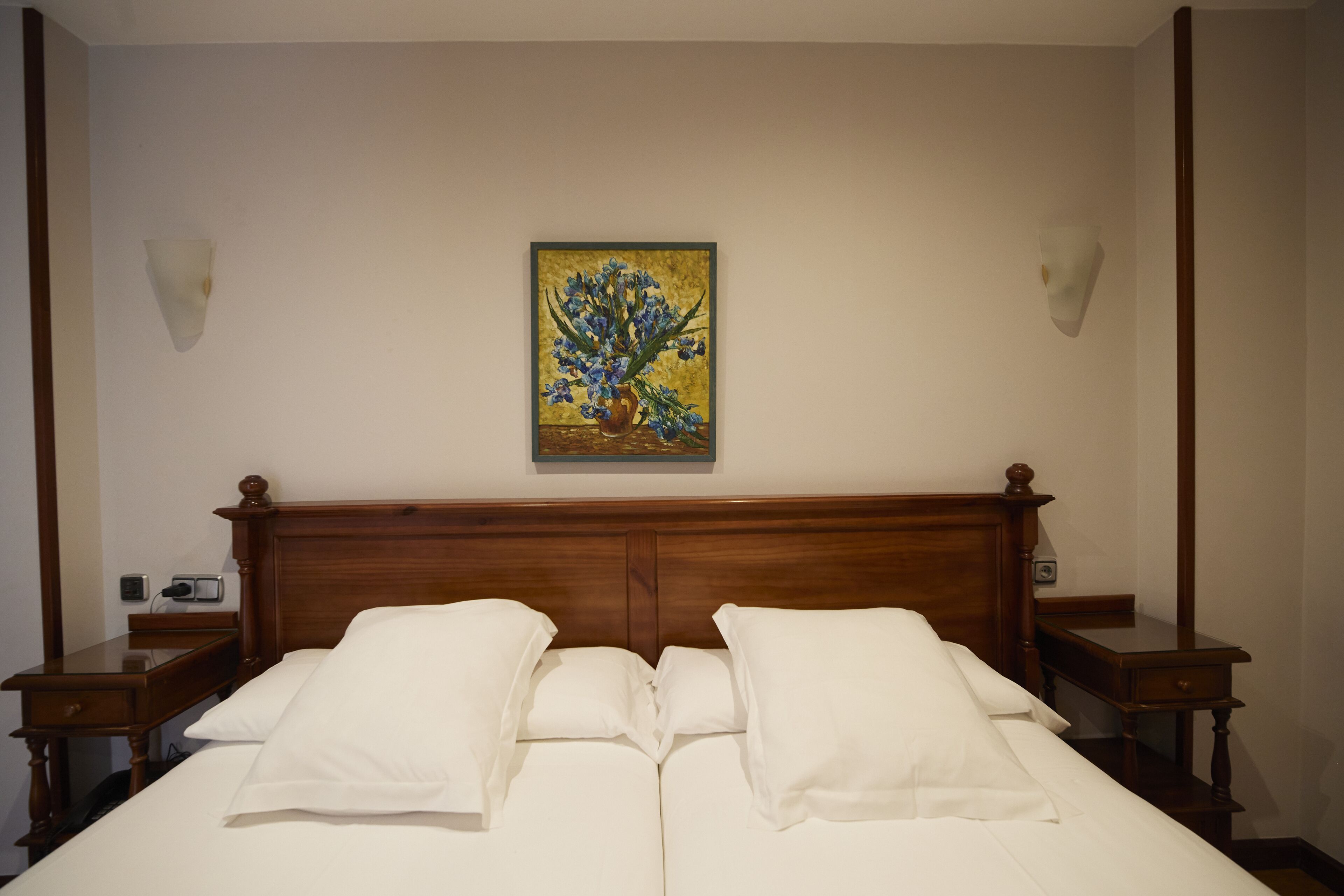 A neatly made bed with white linens and pillows, a wooden headboard, and a painting of flowers on the wall.