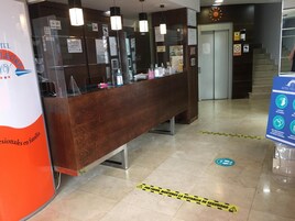 Bancada de recepção de hotel com balcão de madeira, divisória de vidro e sinalização sobre medidas de segurança para a COVID-19.