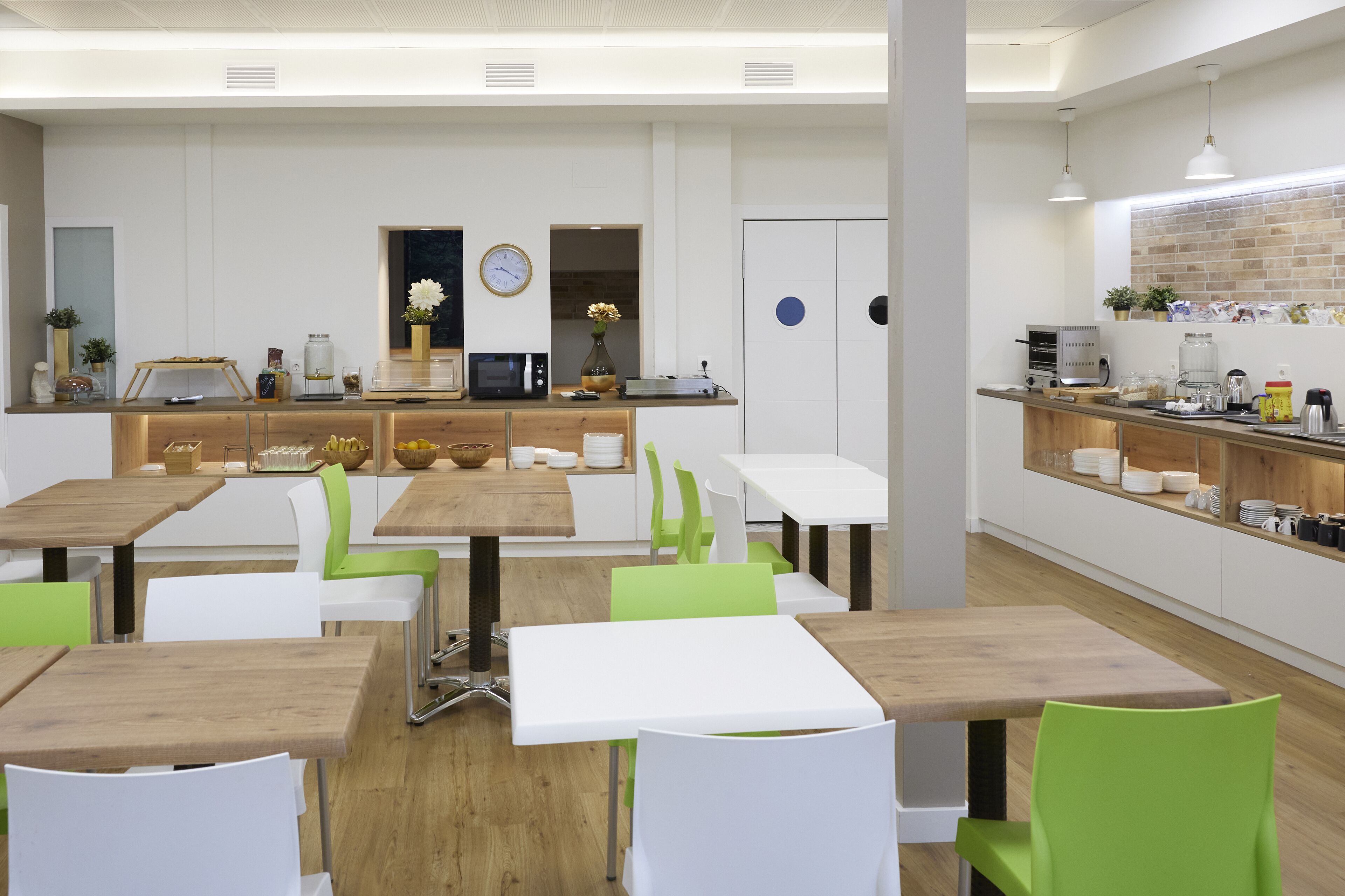 Un moderno caffè con tavoli di legno e sedie verdi, un'area cucina con elettrodomestici e una sveglia appesa al muro.
