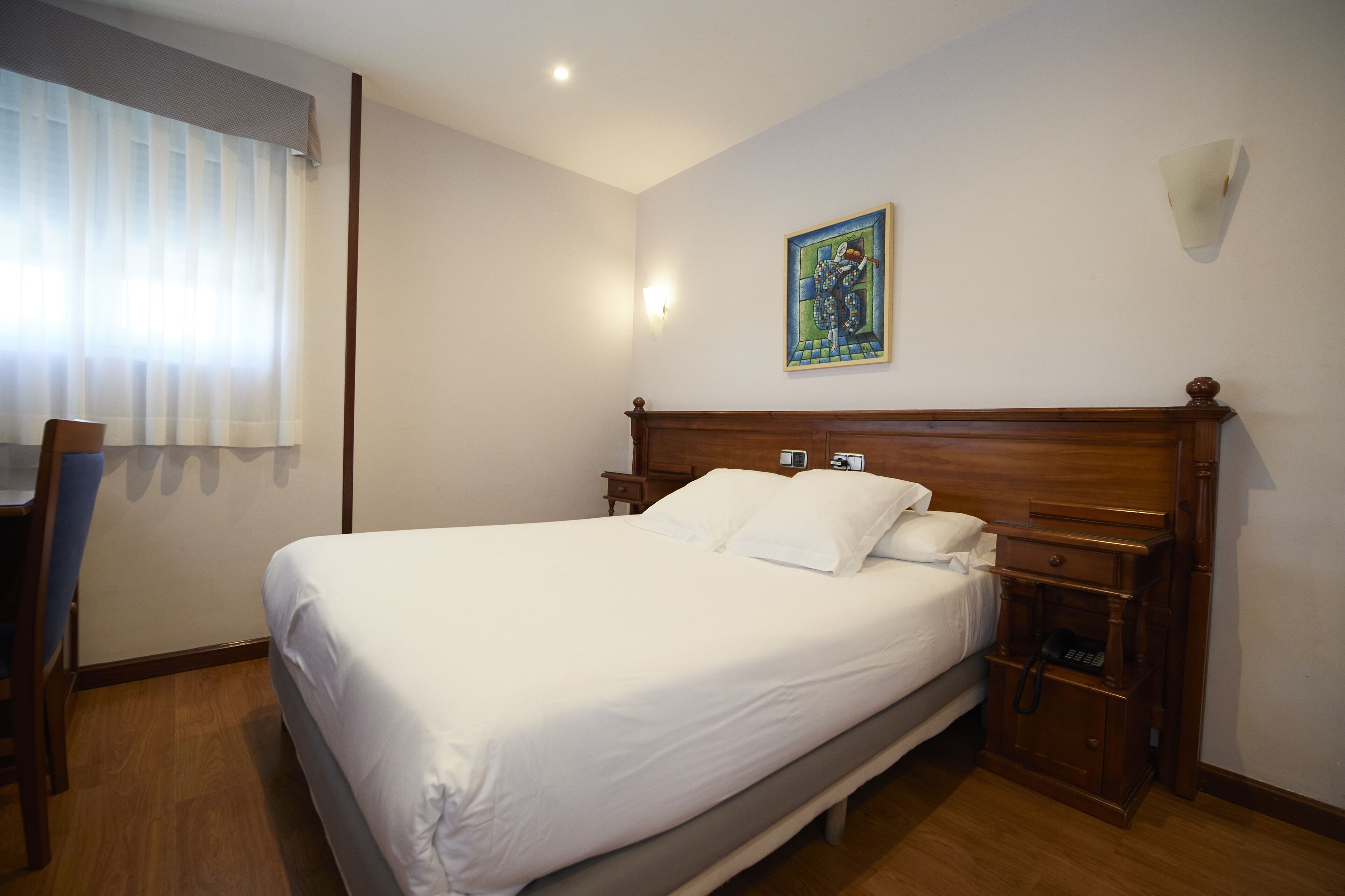 A neatly made bed in a hotel room with a wooden headboard, a bedside table with a phone, and a painting on the wall.