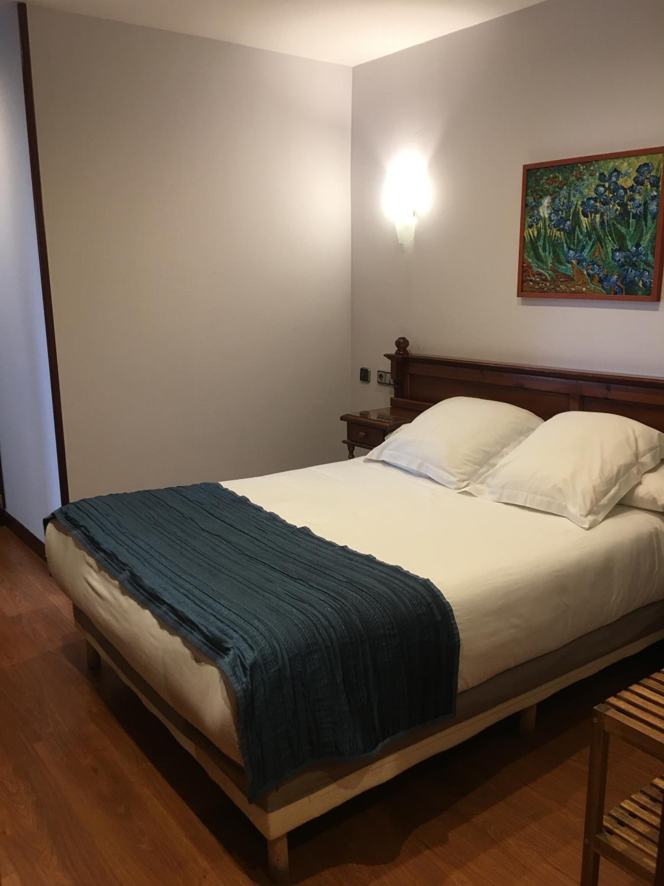 Een net opgemaakt bed met witte lakens en een blauwe deken, een houten hoofdbord, een nachtkastje met een lamp en een schilderij aan de muur.